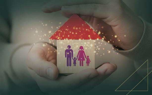 Hands holding a small wooden block house with a red roof. A purple family silhouette, including parents and two children, is on the side of the house. Golden light particles surround the house