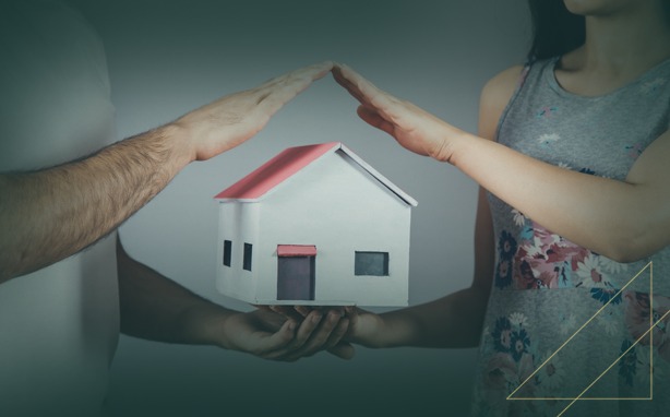 Two people protect a miniature house with their hands, symbolizing home security and care.