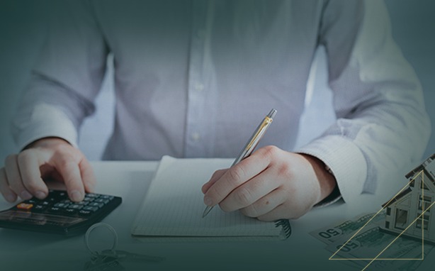 Person calculating and writing at a desk with a calculator, notebook, pen, money, and house model.