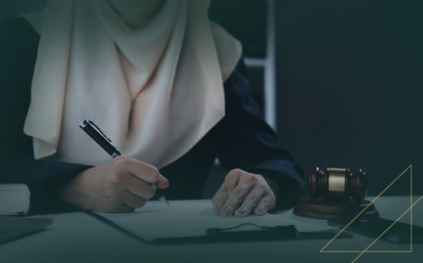 Person in hijab writing at a table with a gavel nearby, gold geometric design in foreground.
