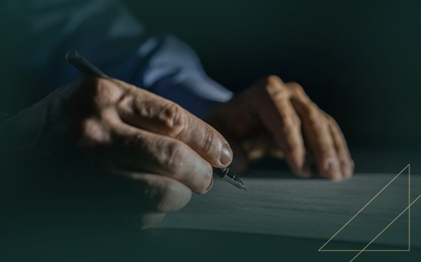 Hand holding a black and silver pen over white paper with a dark, blurred background and gold geometric lines.