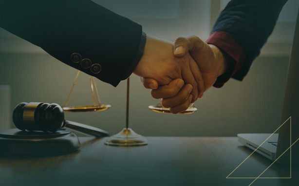 Two people shaking hands over a desk with a scale of justice, gavel, and laptop.