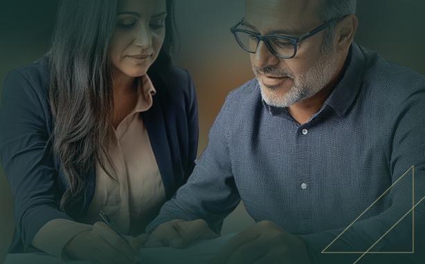 A man with glasses and a beard and a woman with long dark hair review documents together. The woman is writing on a document with a pen, and the man is looking down at the document