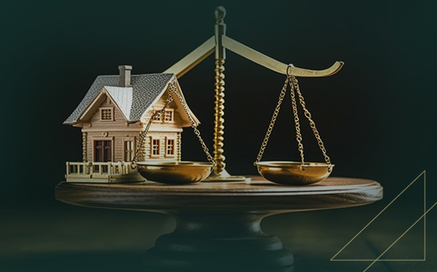 A small wooden house model sits on the left pan of a golden scale, which is balanced on a dark wooden base against a dark background. The right pan of the scale is empty.