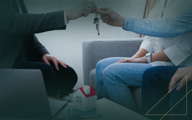 Person in suit hands house keys to another, with a laptop and house model on a table.